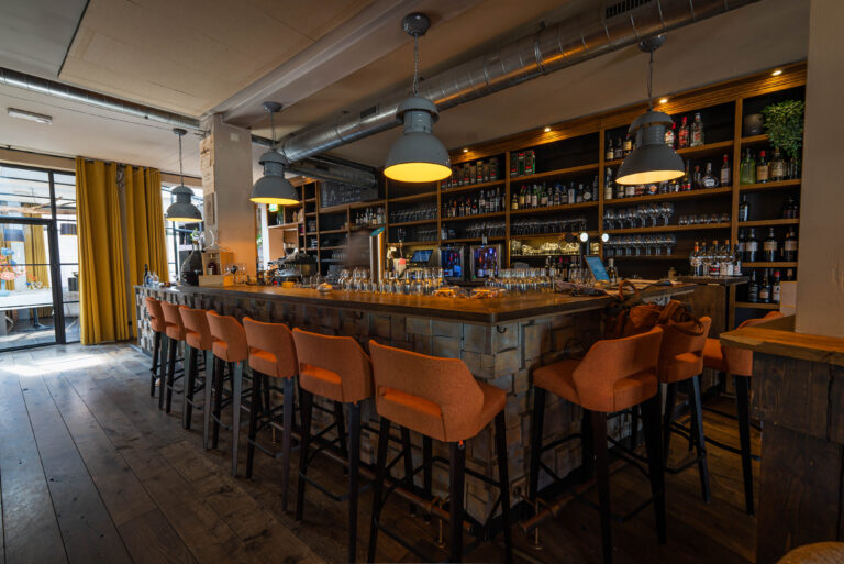 Sfeervolle bar met lange houten toonbank, oranje barkrukken, wand vol flessen en grijze hanglampen.