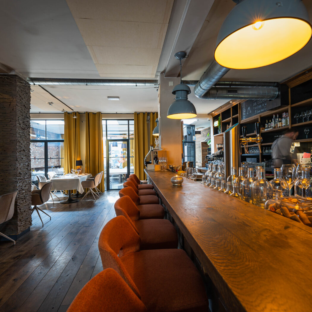 Modern restaurantinterieur met een lange houten bar, oranje krukken, een barman en ramen met gele gordijnen.