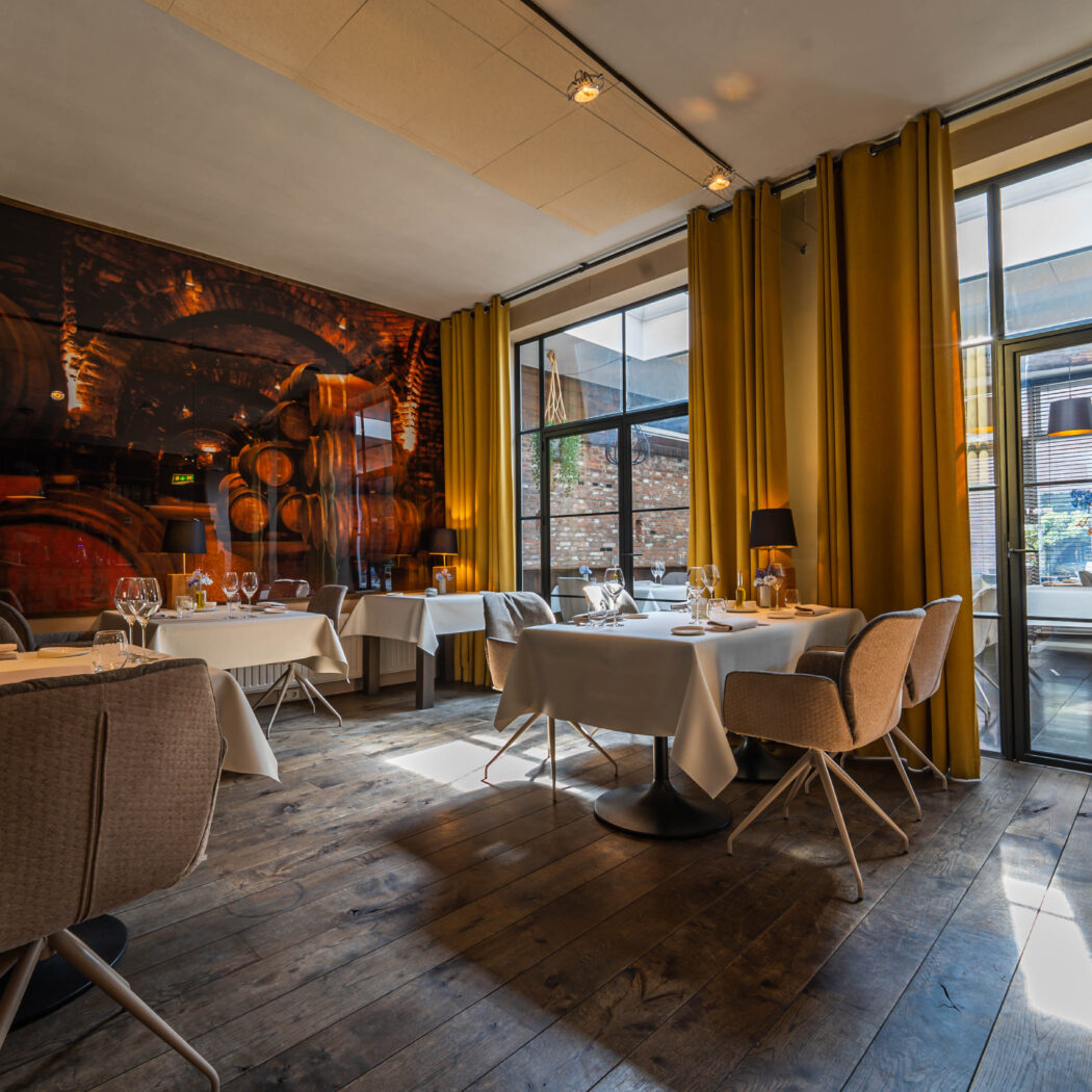 Ruime restaurant eetzaal met houten vloer, wit gedekte tafels, gele gordijnen en wandkunst van wijnvaten, badend in natuurlijk licht.