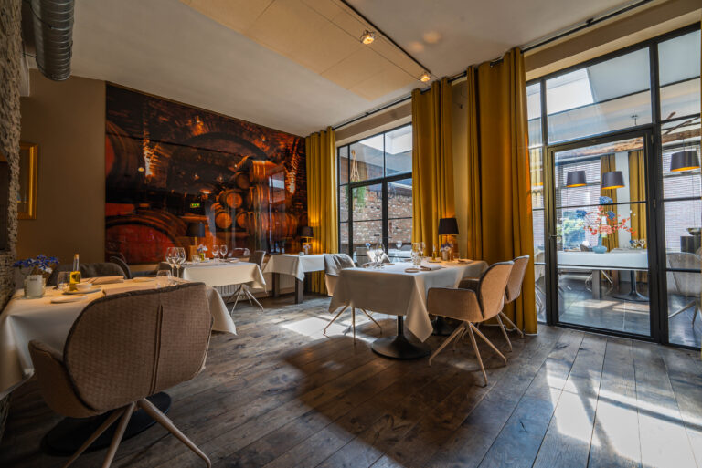 Ruime restaurant eetzaal met houten vloer, wit gedekte tafels, gele gordijnen en wandkunst van wijnvaten, badend in natuurlijk licht.