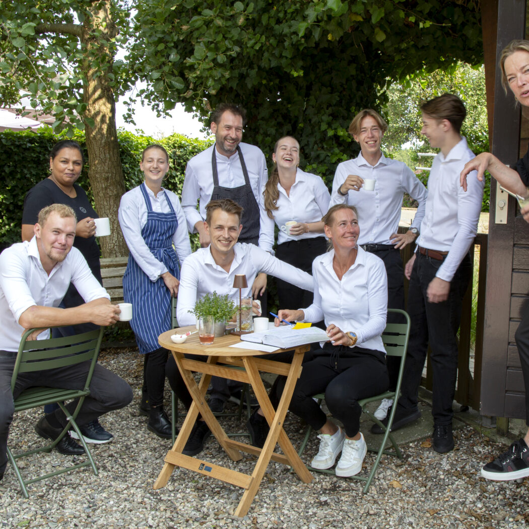 Vrolijk personeelsteam met witte overhemden en schorten zit en staat samen op een zonnig buitenterras.