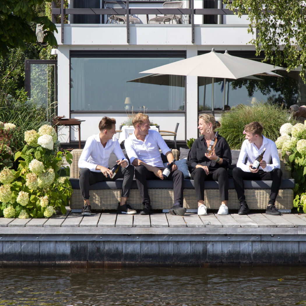 Vier mensen zitten lachend op een rieten bank aan een houten vlonder bij het water, met drankflessen en hortensias.