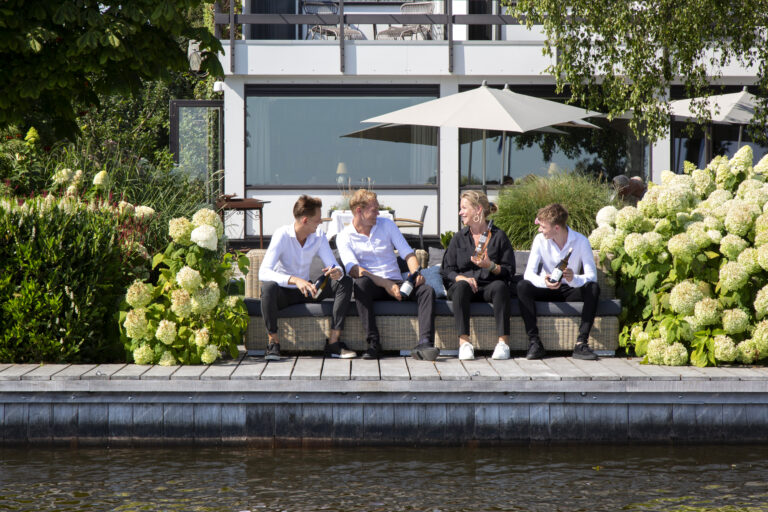 Vier mensen zitten lachend op een rieten bank aan een houten vlonder bij het water, met drankflessen en hortensias.