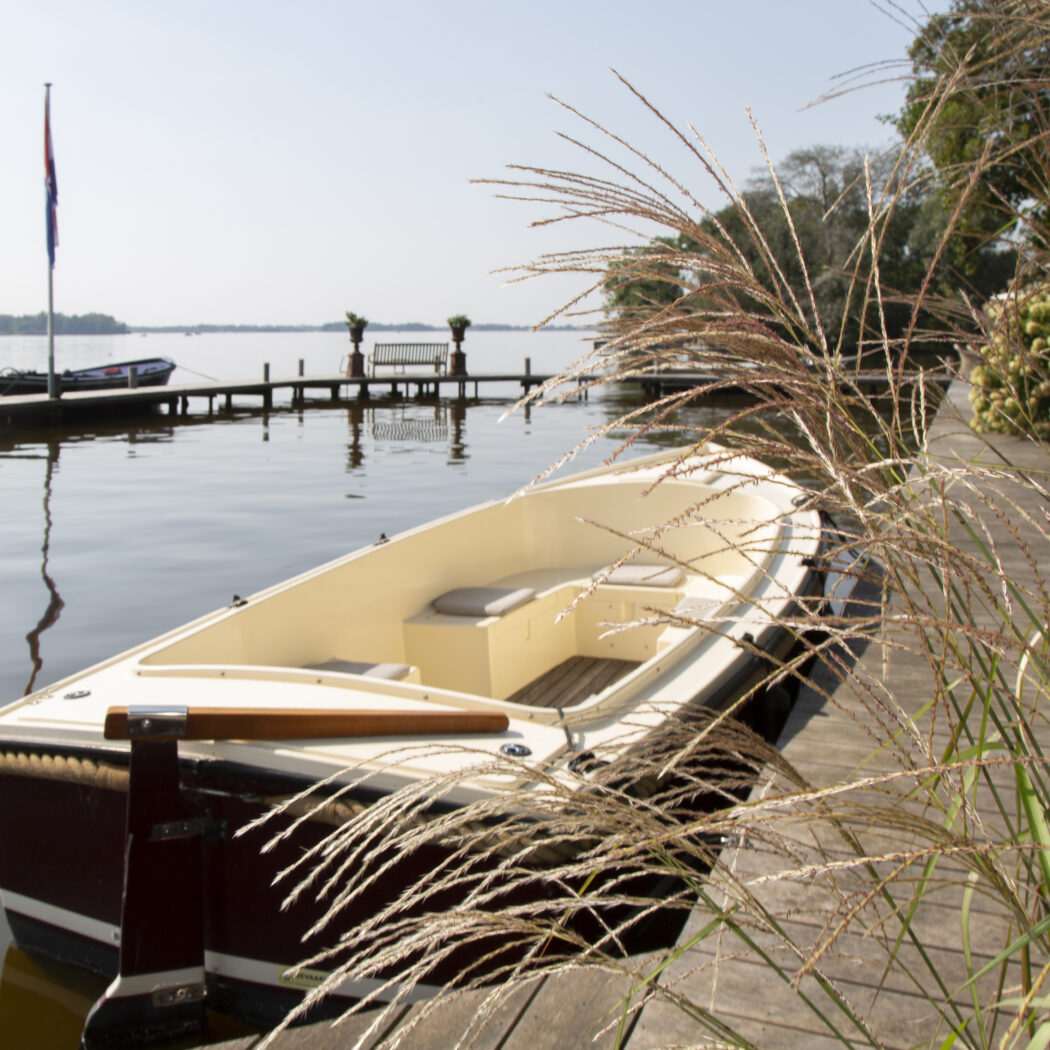 Crème-witte sloep met donkerrode romp ligt aan een houten steiger op rustig water, met siergras.