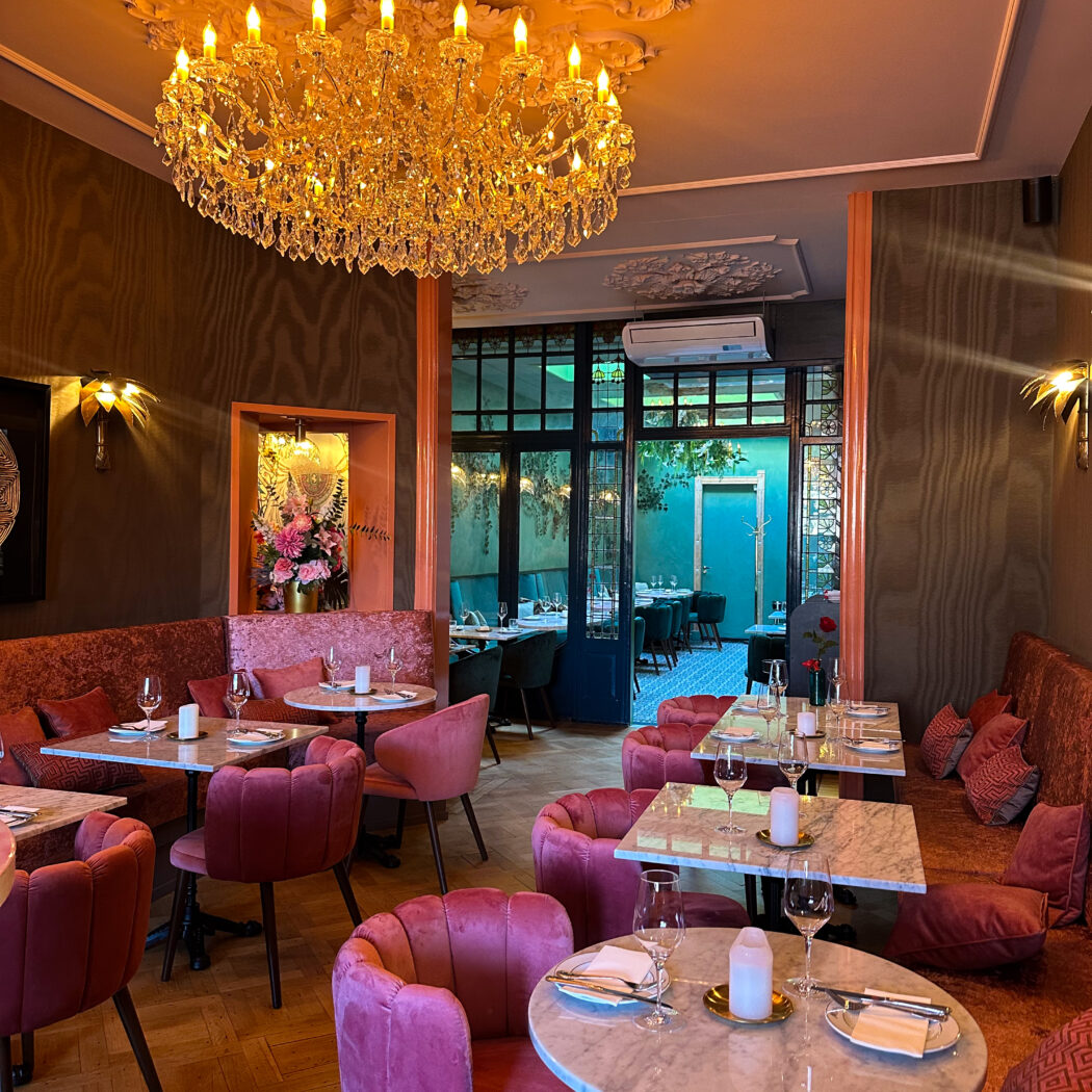 Elegant restaurantinterieur met een grote kristallen kroonluchter, roze fluwelen stoelen, marmeren tafels en houten vloer.