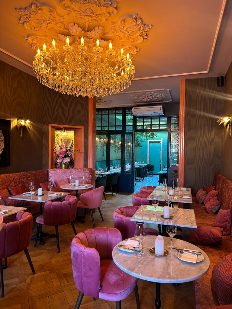 Elegant restaurantinterieur met een grote kristallen kroonluchter, roze fluwelen stoelen, marmeren tafels en houten vloer.