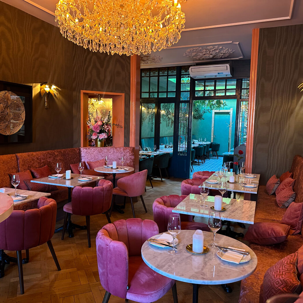 Sfeervol restaurant met oranje fluwelen banken, roze stoelen en gedekte marmeren tafels onder een kristallen kroonluchter.