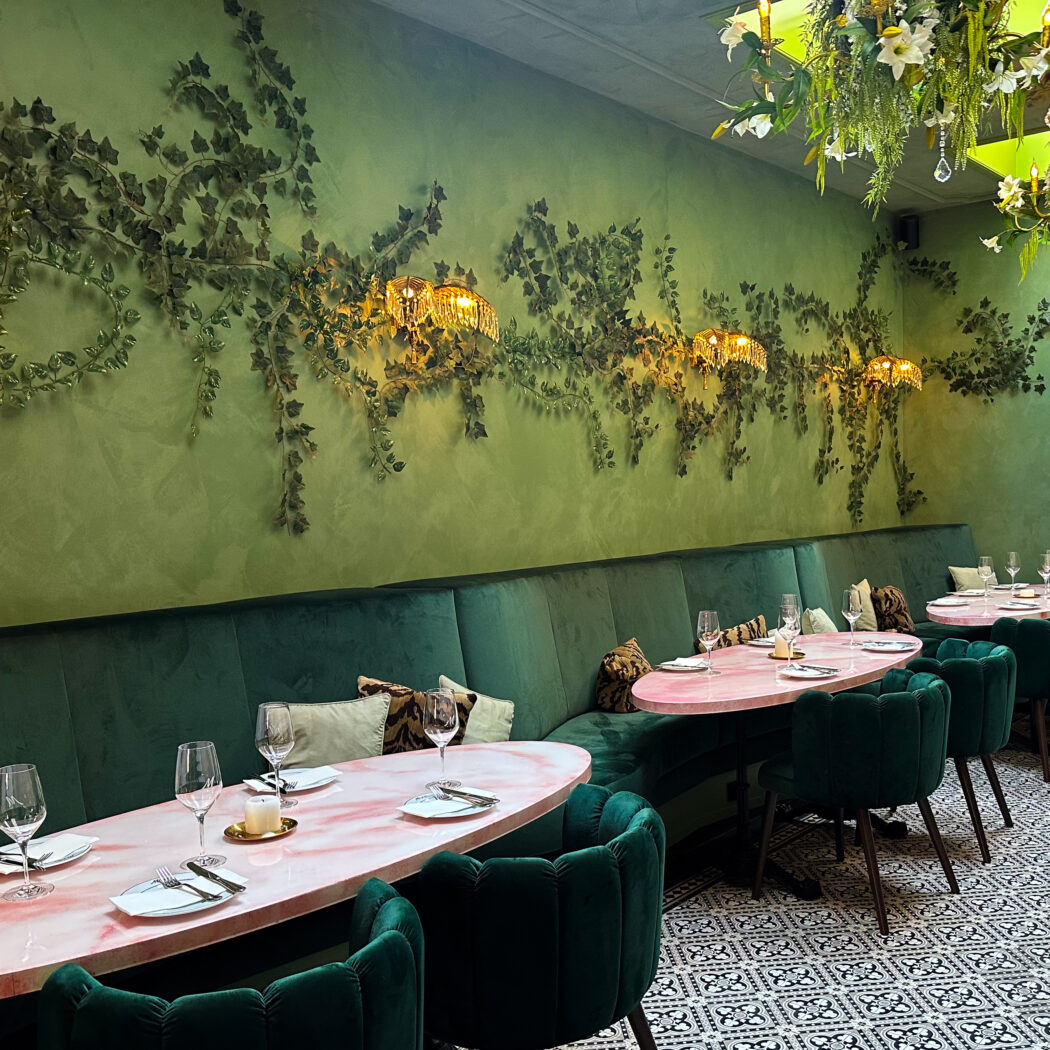 Sfeervol restaurant met groene muren, klimopdecoraties, gouden lampen, roze marmeren tafels en smaragdgroene fluwelen stoelen.