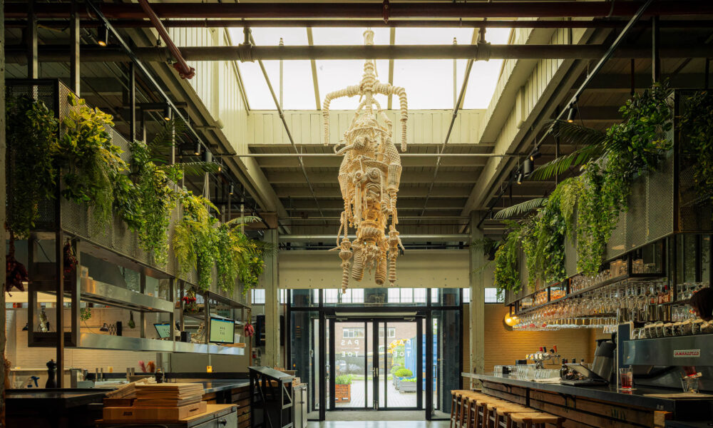 Large, industrial restaurant interior with unique rope sculpture, hanging plants, bar and the text PAS TOE.