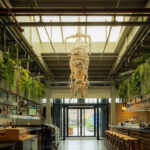 Large, industrial restaurant interior with unique rope sculpture, hanging plants, bar and the text PAS TOE.