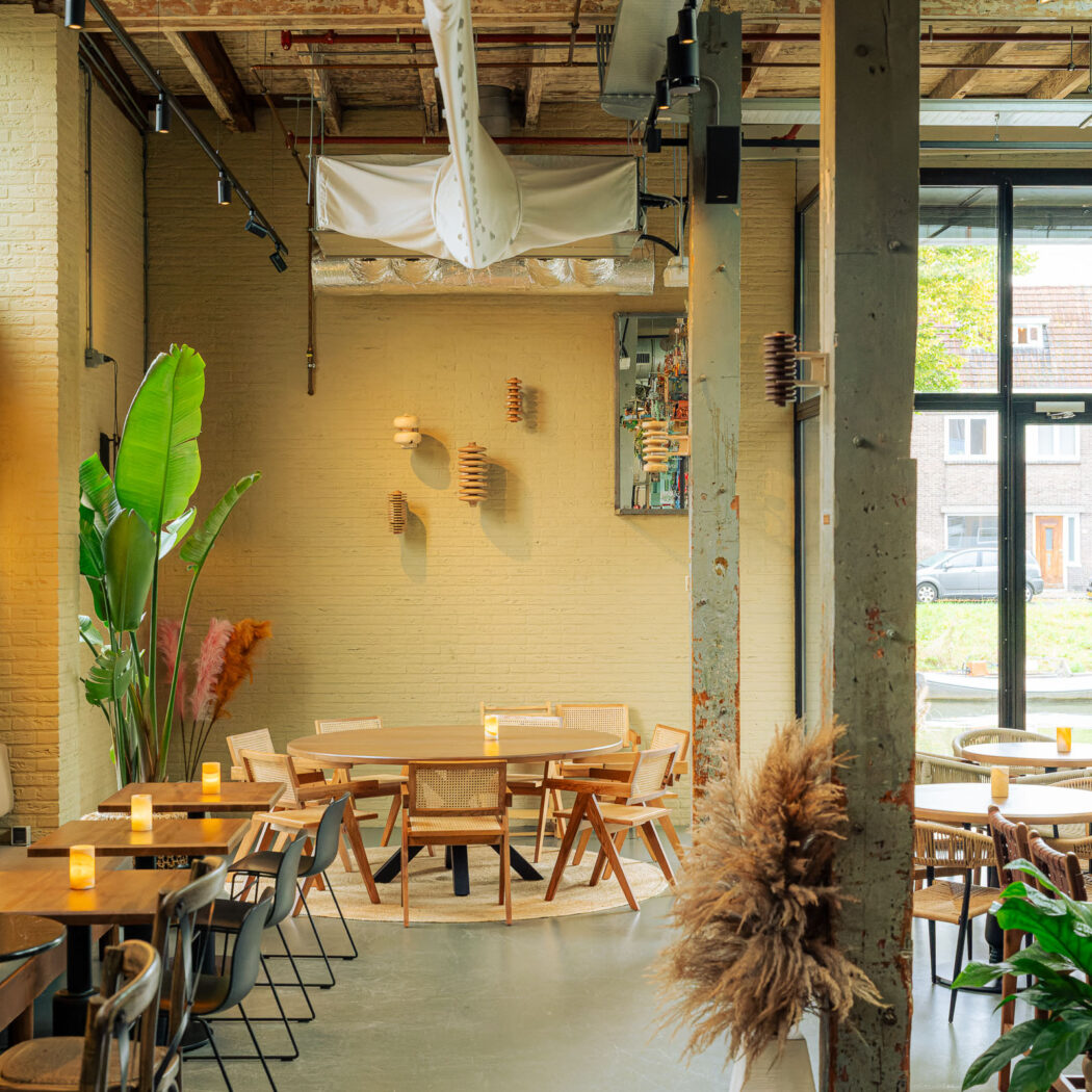 Licht restaurant met houten tafels, veel planten, industriële lampen aan gele bakstenen muren en uitzicht op een gracht.