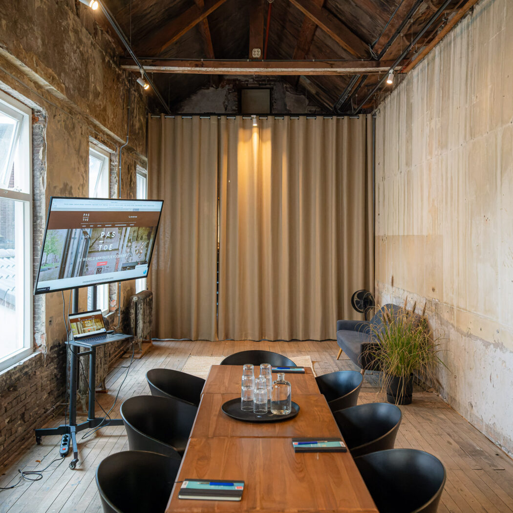Industriële vergaderzaal met lange houten tafel, zwarte stoelen, scherm met PAS TOE website en grote ramen.