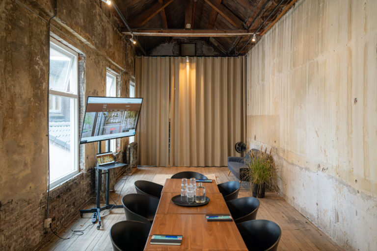 Industriële vergaderzaal met lange houten tafel, zwarte stoelen, scherm met PAS TOE website en grote ramen.