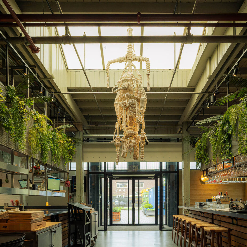 Groot, industrieel restaurantinterieur met unieke touwsculptuur, hangplanten, bar en de tekst PAS TOE.