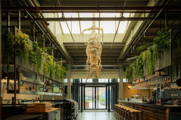 Groot, industrieel restaurantinterieur met unieke touwsculptuur, hangplanten, bar en de tekst PAS TOE.