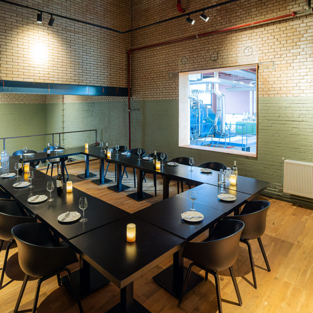 U-vormige zwarte tafel met kaarsen en gedekt servies in een zaal met beige bakstenen muren en uitzicht op blauwe machines.