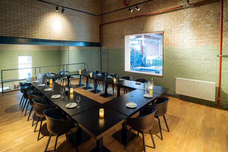 U-vormige zwarte tafel met kaarsen en gedekt servies in een zaal met beige bakstenen muren en uitzicht op blauwe machines.