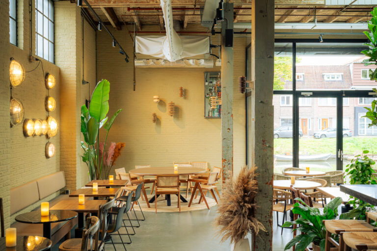 Licht restaurant met houten tafels, veel planten, industriële lampen aan gele bakstenen muren en uitzicht op een gracht.