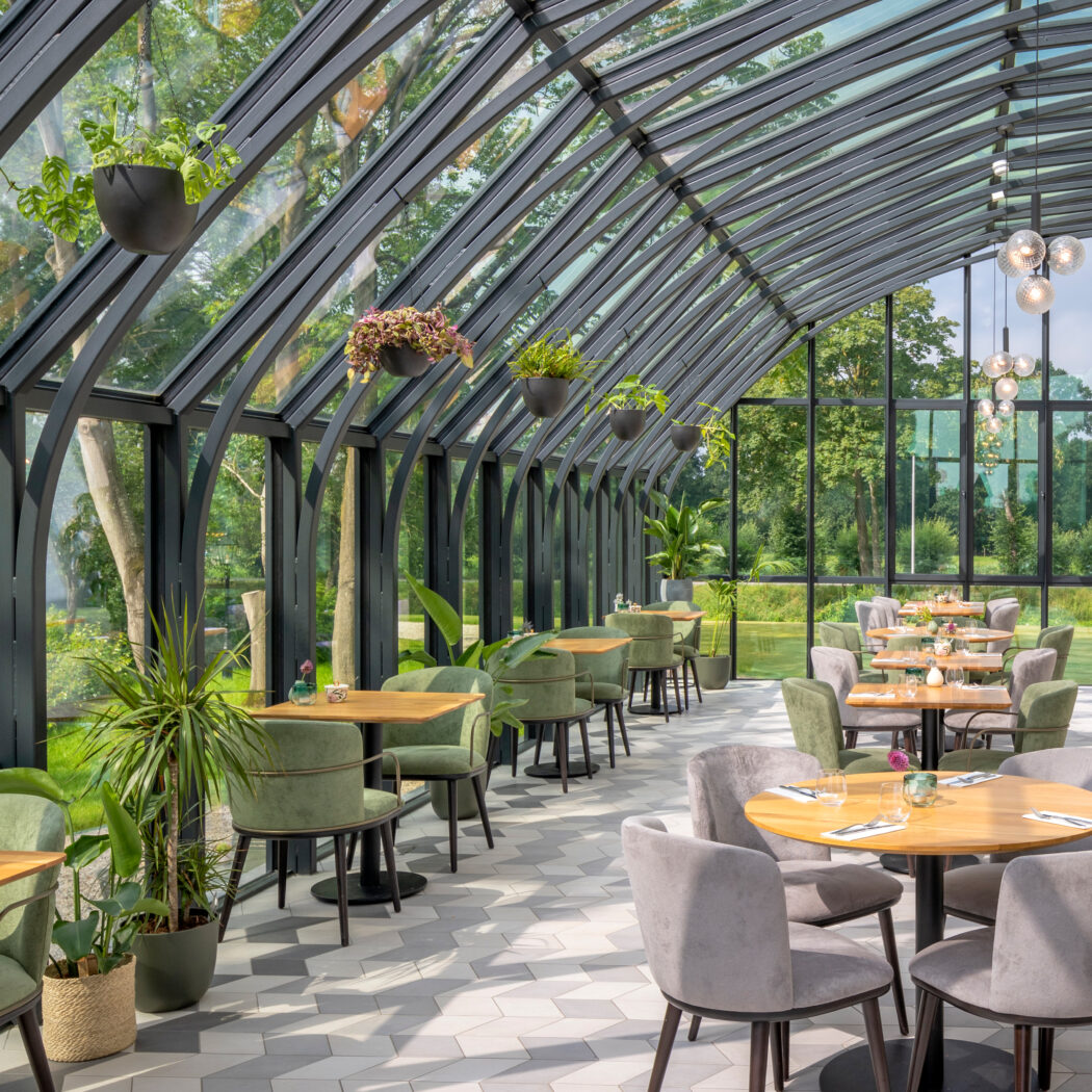Modern serre-restaurant met glazen dak, groene en grijze stoelen aan houten tafels, en weelderig groen.