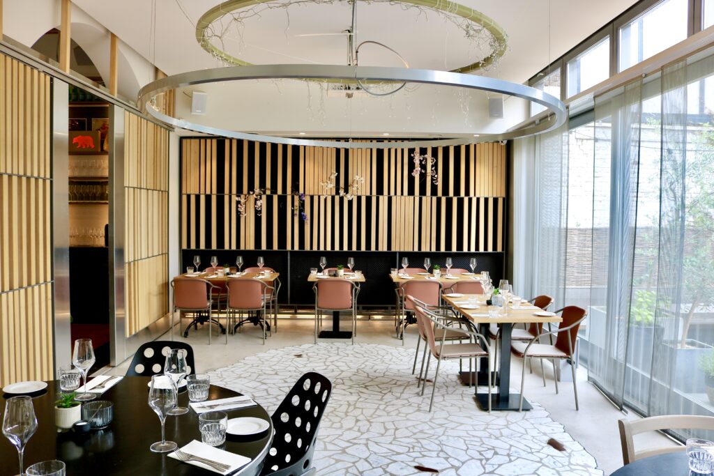 Modern restaurant interieur met houten wandpanelen, gedekte tafels, roze stoelen en grote ronde plafondverlichting.