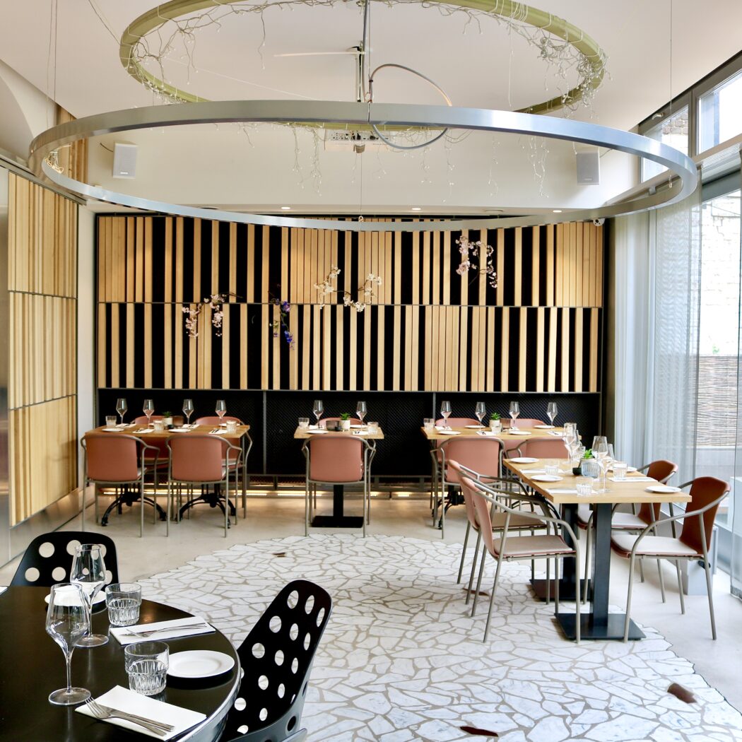 Modern restaurant interieur met houten wandpanelen, gedekte tafels, roze stoelen en grote ronde plafondverlichting.
