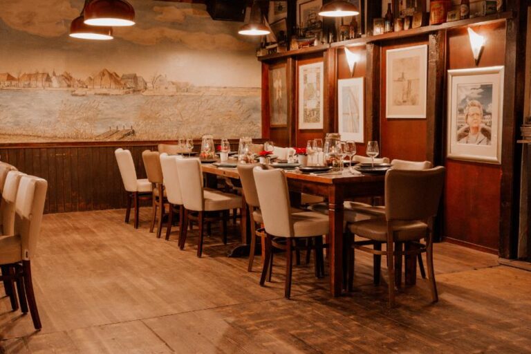 Sfeervolle eetzaal met lange gedekte houten tafel, lichte stoelen en muurschildering van een Hollands landschap.