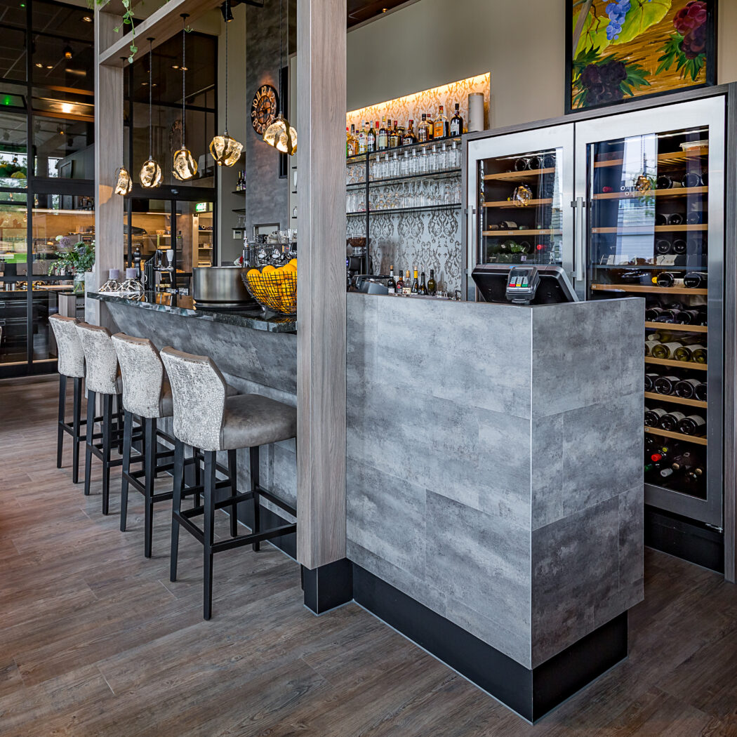 Modern restaurantinterieur met grijze stenen bar, vijf barkrukken en twee wijnkoelkasten.