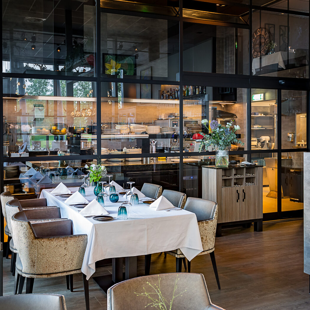 Modern restaurantinterieur met gedekte eettafel, beige stoelen en glazen wand naar de keuken.