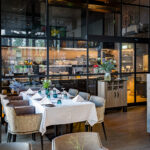 Modern restaurantinterieur met gedekte eettafel, beige stoelen en glazen wand naar de keuken.