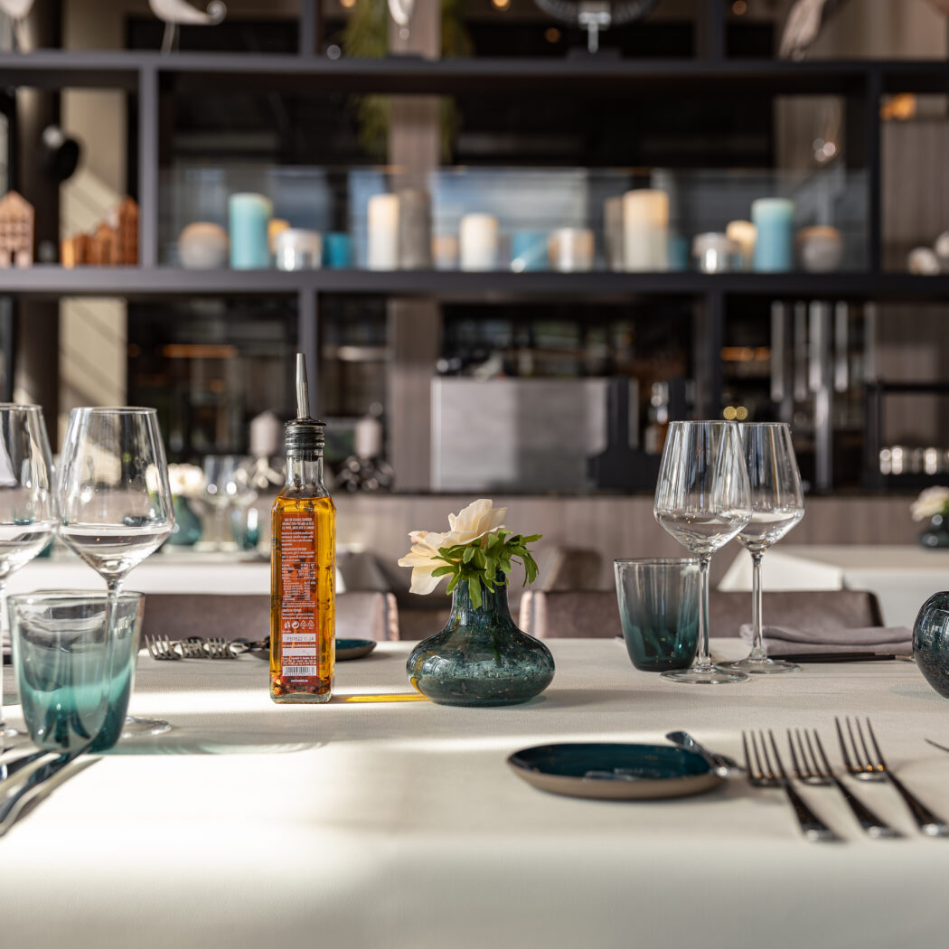 Elegant gedekte restauranttafel met wijnglazen, bestek, olijfolie, een witte bloem en blauwgroene decoratie.