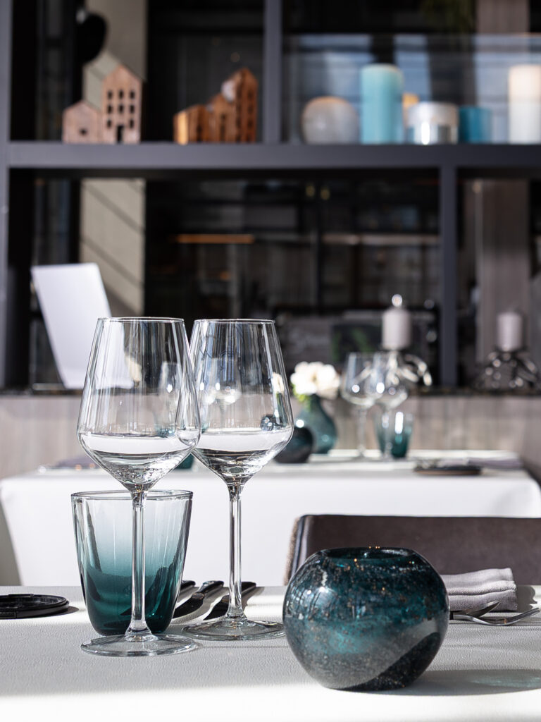 Gedekte tafel in een restaurant met heldere wijnglazen, een groenblauw drinkglas en een decoratieve bol.
