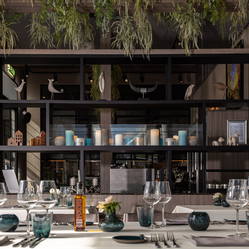 Elegant gedekte restauranttafel met wijnglazen en olijfolie, voor een schap vol kaarsen, ornamenten en hangplanten.