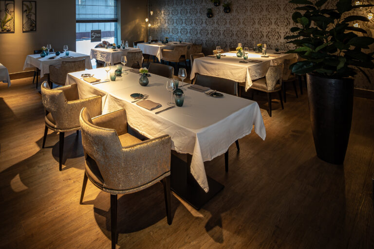 Elegant restaurantinterieur met wit gedekte tafels, sfeervolle verlichting en comfortabele goudkleurige stoelen.