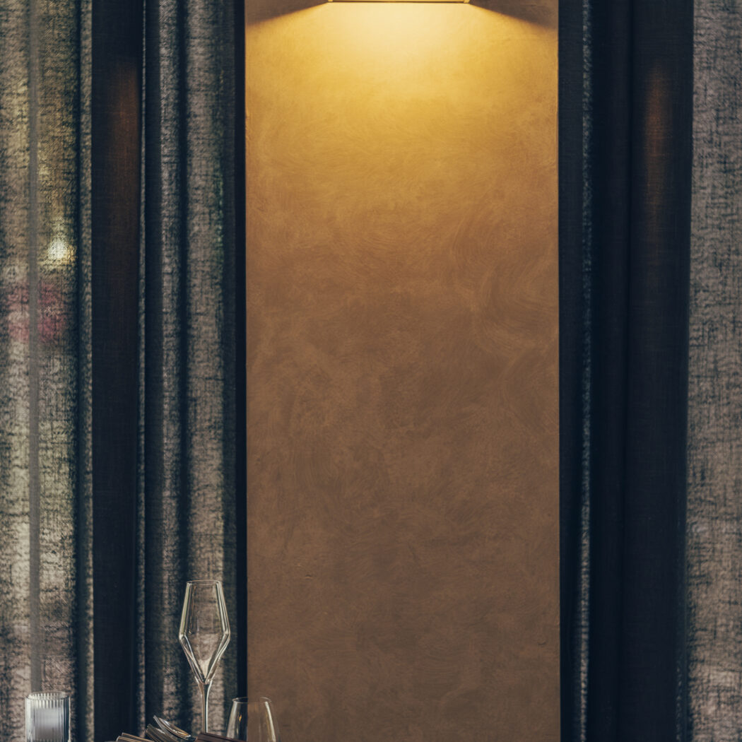 Sfeervolle restauranttafel met glaswerk, houten stoel, en een wandlamp op een textuurmuur met donkere gordijnen.
