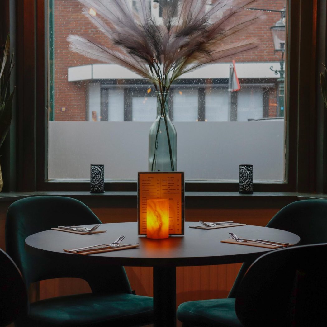 Gezellige ronde tafel voor vier met teal fluwelen stoelen, brandende kaars, en decoratieve veren en planten bij een raam.