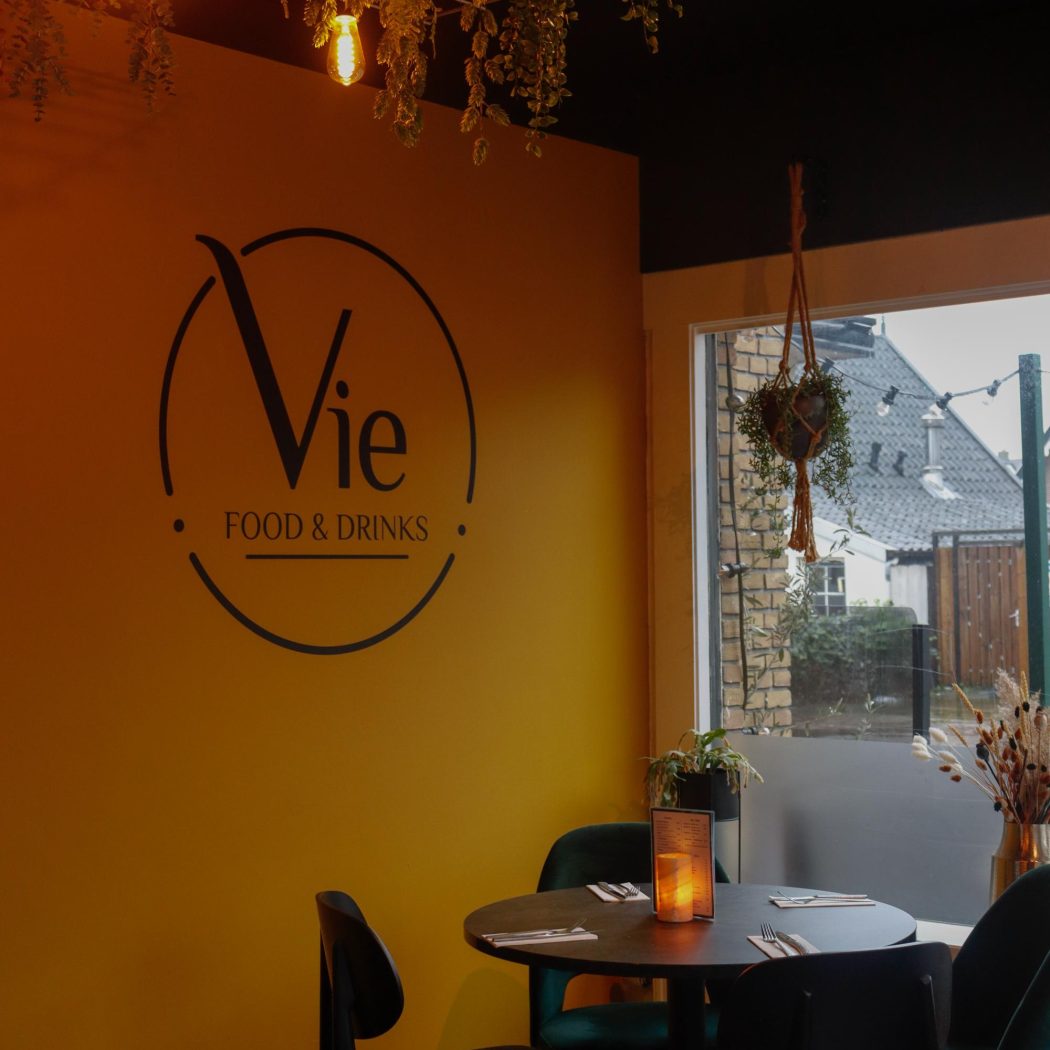 Sfeervol café interieur met geel Vie FOOD & DRINKS logo, donkere tafel, groene stoelen en hangplanten.