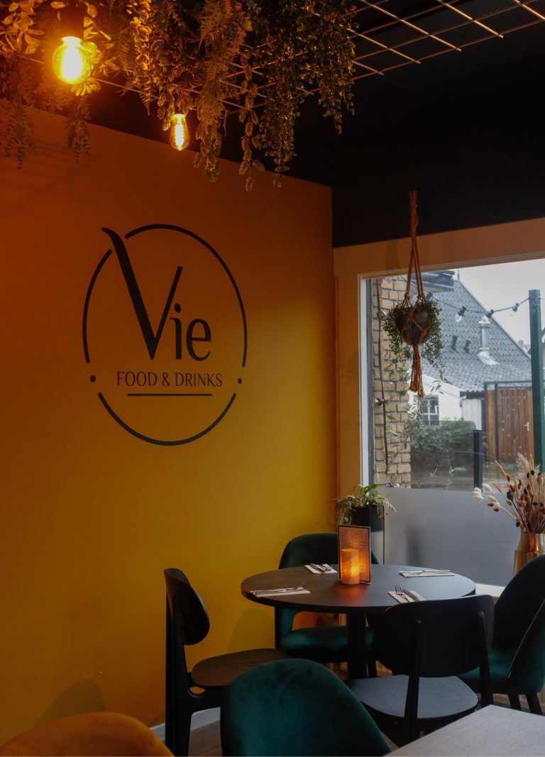Sfeervol café interieur met geel Vie FOOD & DRINKS logo, donkere tafel, groene stoelen en hangplanten.