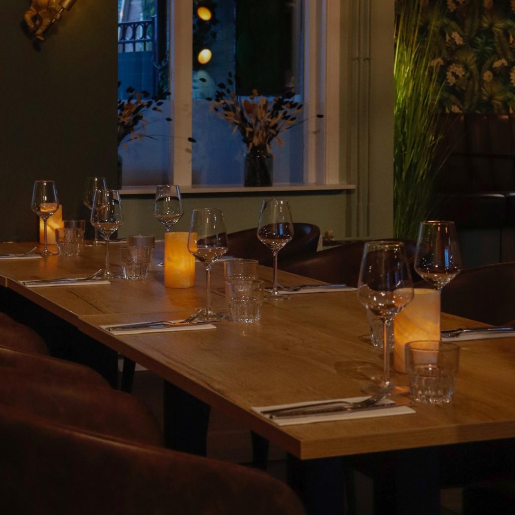 Lange houten restauranttafel met glazen, bestek, sfeervolle kaarsen en bruine leren stoelen.