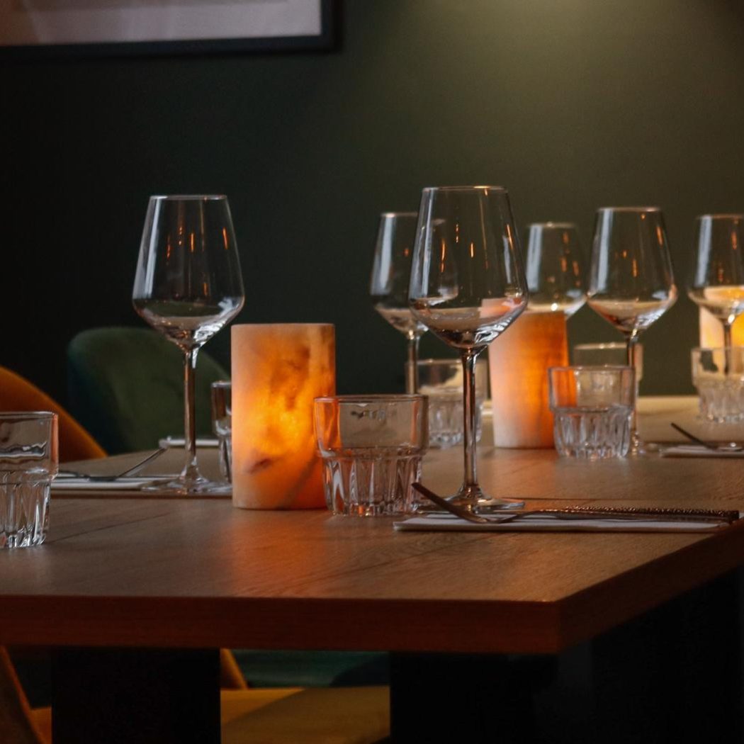 Lange houten restauranttafel gedekt met glazen en bestek, verlicht door sfeervolle zoutlampen, met groene en oranje stoelen.
