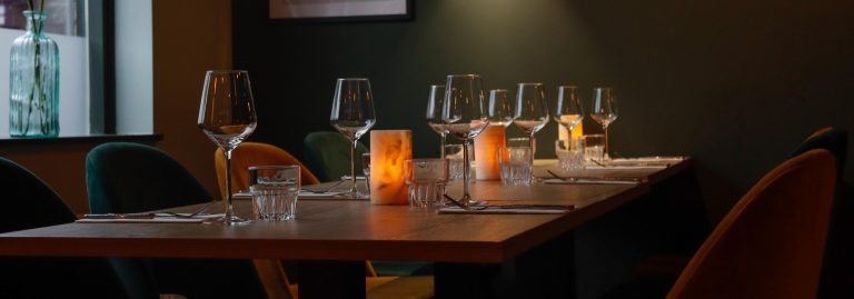 Lange houten restauranttafel gedekt met glazen en bestek, verlicht door sfeervolle zoutlampen, met groene en oranje stoelen.