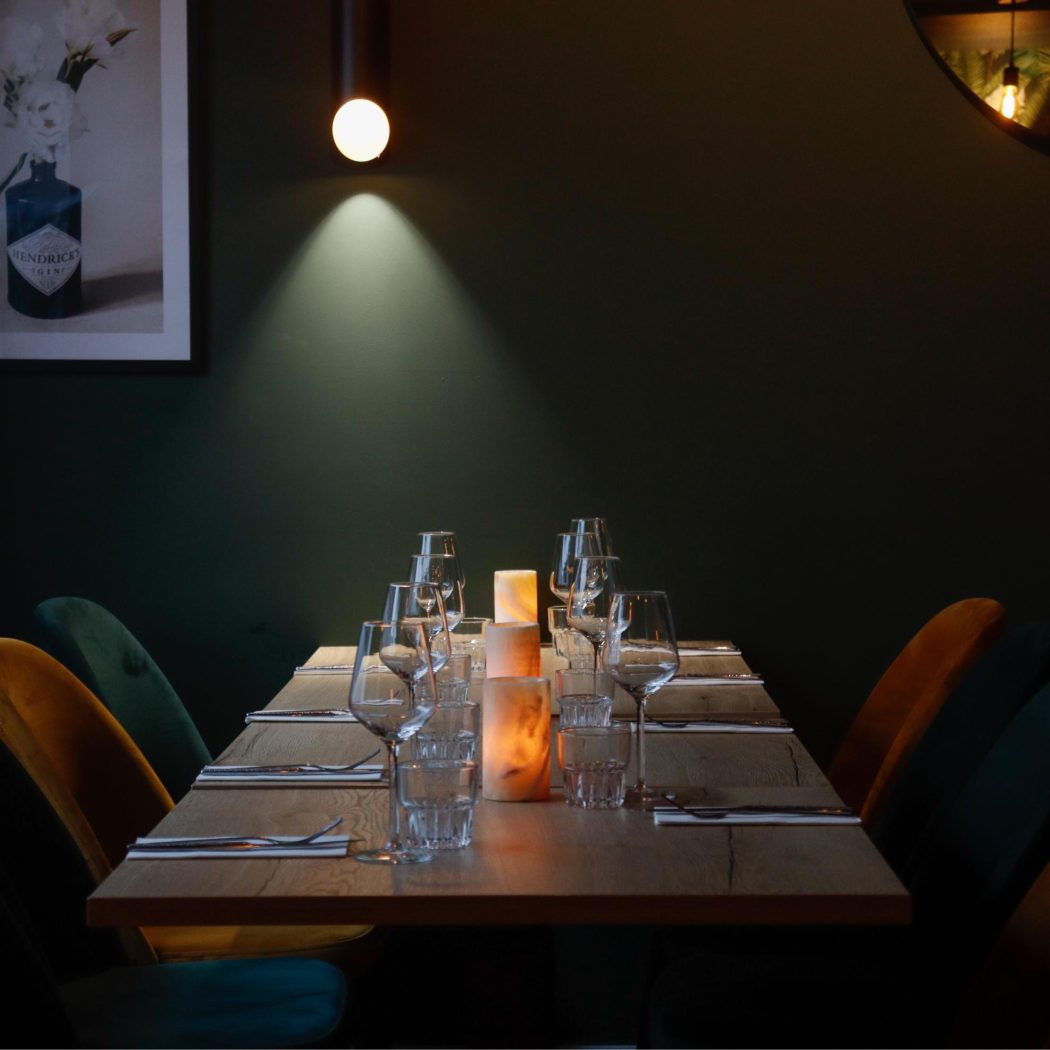 Gezellige restauranttafel met glazen, zoutlampen en teal/okergele velours stoelen onder een spot, bij Hendricks Gin poster.