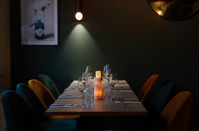 Gezellige restauranttafel met glazen, zoutlampen en teal/okergele velours stoelen onder een spot, bij Hendricks Gin poster.