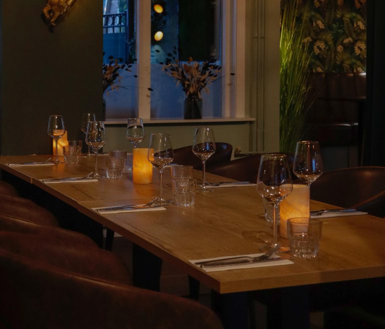 Lange houten restauranttafel met glazen, bestek, sfeervolle kaarsen en bruine leren stoelen.