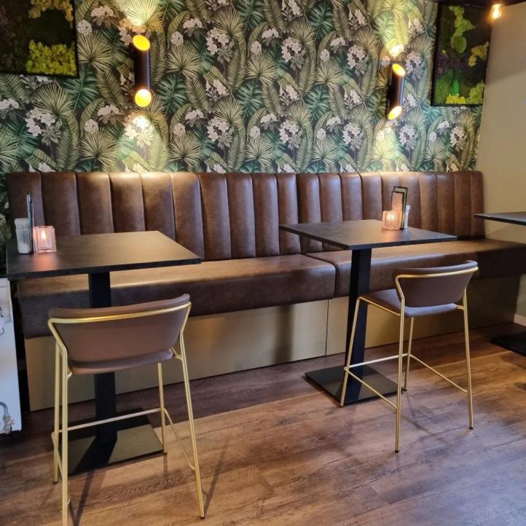 Gezellige zithoek met bruine bank, zwarte tafels en gouden barkrukken tegen een muur met tropisch bladbehang.