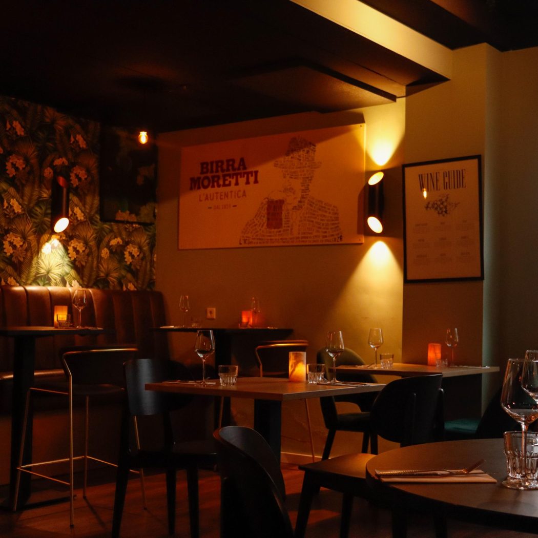 Gezellig restaurantinterieur met sfeervolle verlichting, tafels met wijnglazen en kaarsen, en een BIRRA MORETTI poster.