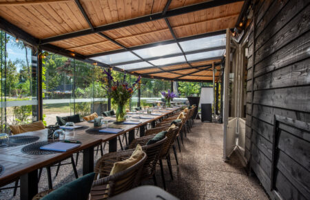 Feestelijk gedekte tafel op een overdekt terras met houten plafond en glazen wanden, omgeven door groen.