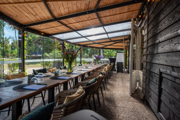 Feestelijk gedekte tafel op een overdekt terras met houten plafond en glazen wanden, omgeven door groen.