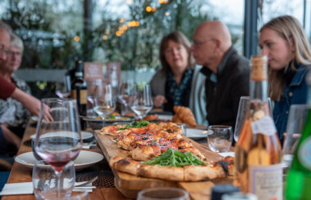 Lange houten plank met gesneden pizza, rucola en olijven, op tafel met Cuvée Prestige rosé en gasten.