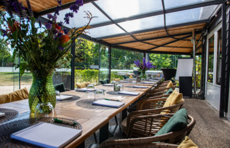 Gedekte houten tafel in een lichte serre met rieten stoelen en bloemdecoratie, uitzicht op tuin.