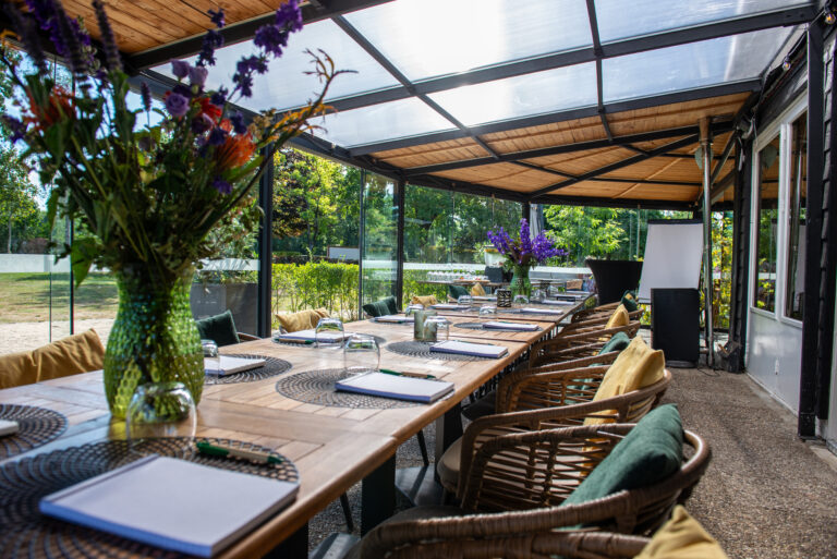 Gedekte houten tafel in een lichte serre met rieten stoelen en bloemdecoratie, uitzicht op tuin.